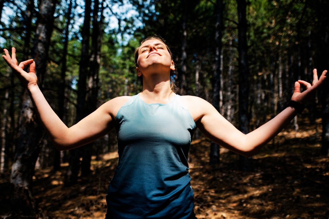 Person outdoors in a forest, basking in the sunlight with eyes closed and arms raised in a peaceful, meditative pose, embodying vitality and energy.