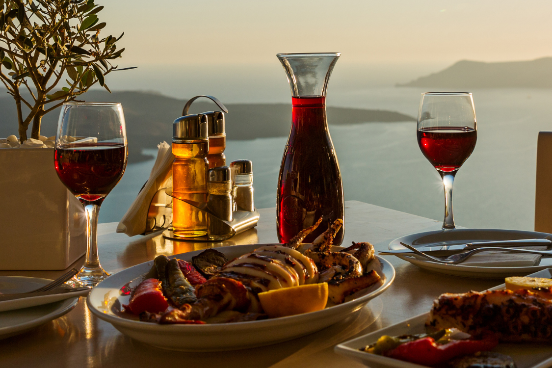 two glasses of wine and a glass bottle of red wine at the dinner table with healthy Mediterranean food and condiments, with the blue sea, blue sky and mountains in the background, at a luxury restaurant
