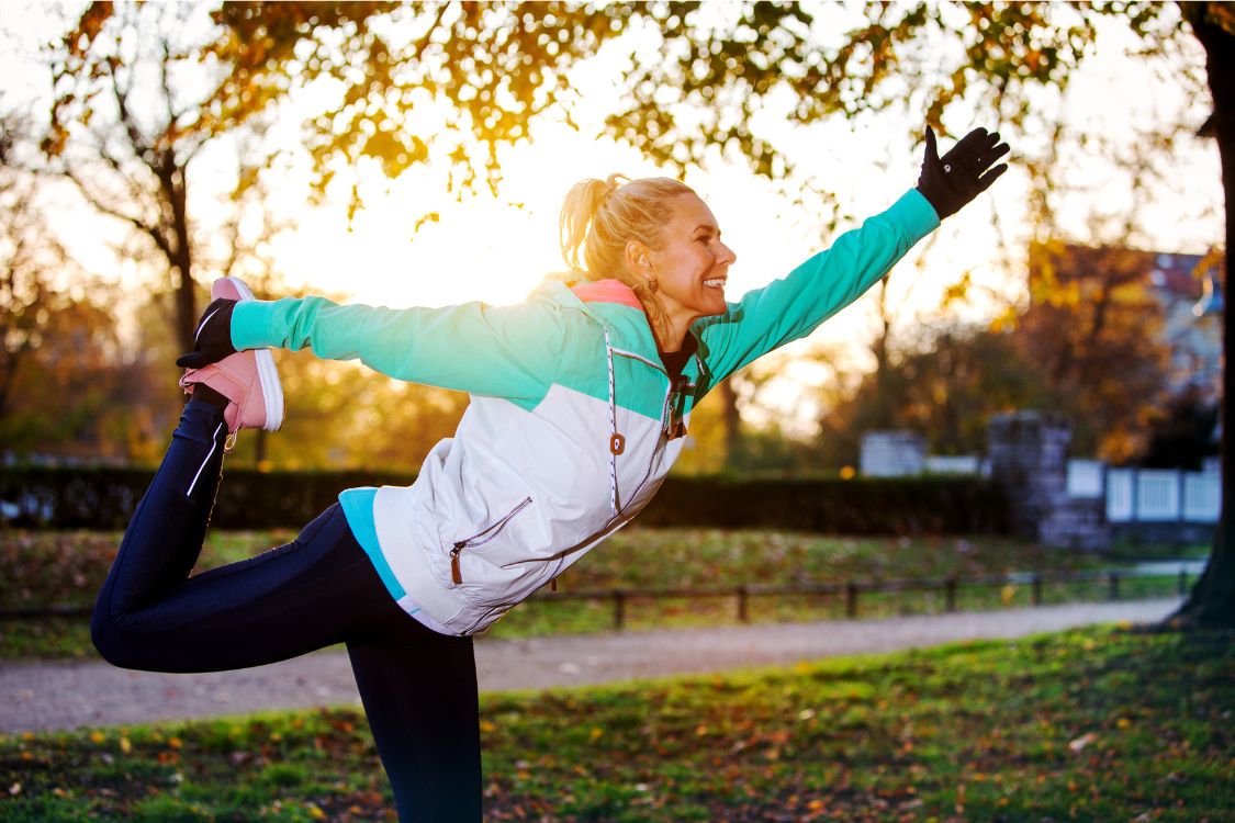 Image of a middle-aged woman exercising outdoors in the morning wearing a green and white jacket stretching and doing physical activity to combat menopausal fatigue